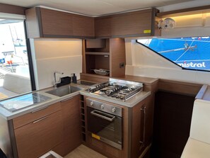 A compact kitchen with a gas stove, oven, and sink, wooden cabinets, and a window with a view of a blue sports field. - Sun-Kissed Sailing Adventure in Islas Sarónicas (Alimos)