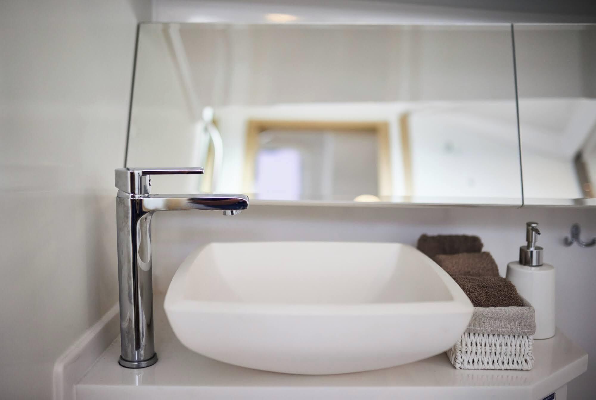 Une salle de bain avec un lavabo blanc, un robinet chromé et un miroir qui reflète un lit et une table de chevet.