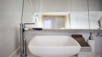 Une salle de bain avec un lavabo blanc, un robinet chromé et un miroir qui reflète un lit et une table de chevet.
