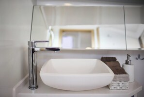 Un baño con lavamanos blanco, grifo cromado y un espejo que refleja una cama y una mesita de noche.