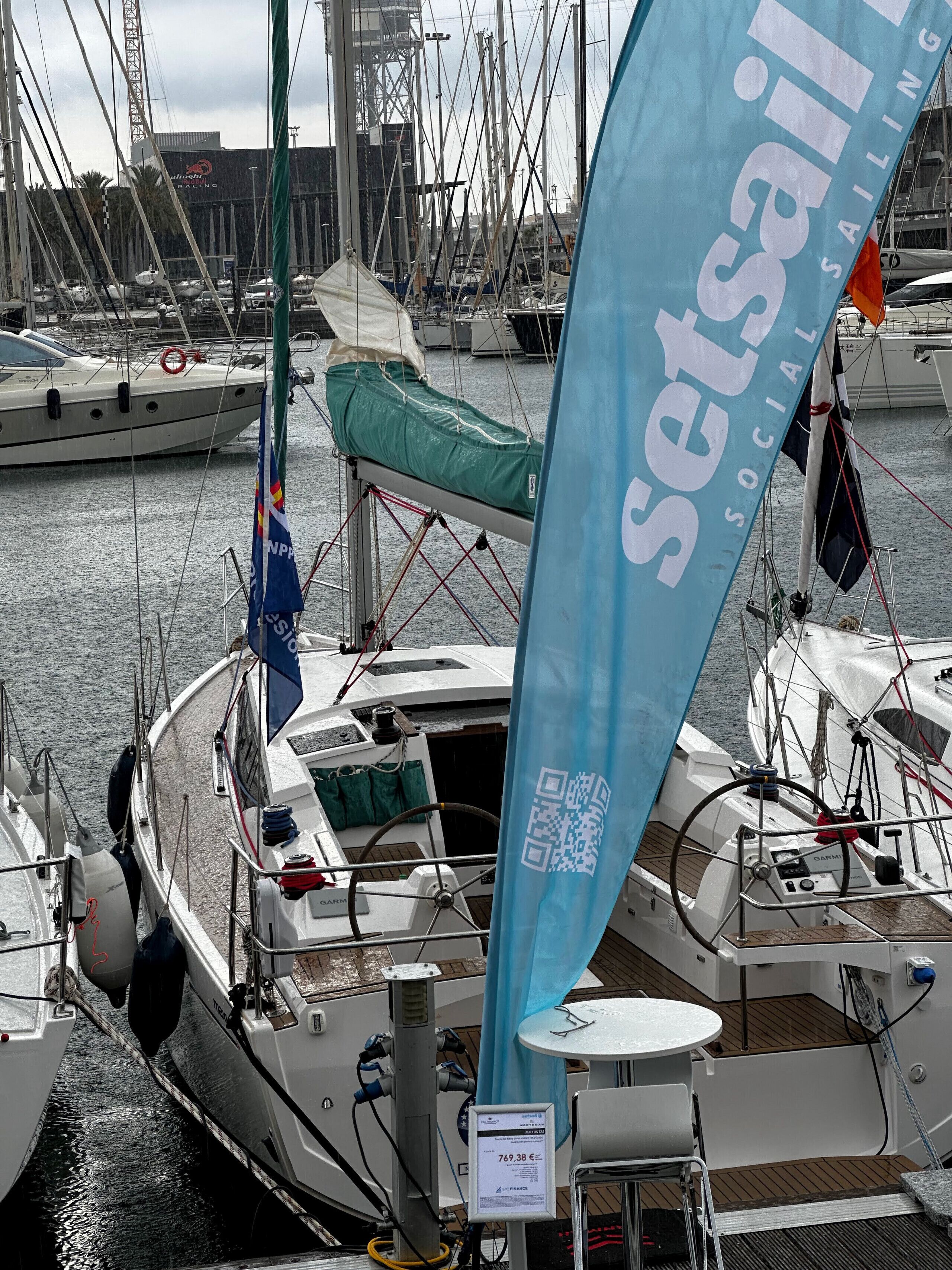 Un port de plaisance où sont amarrés de nombreux voiliers, un code QR sur un drapeau bleu et une table avec une chaise sur le pont.