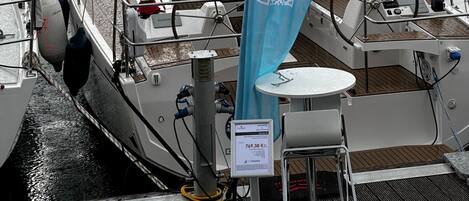 Un port de plaisance où sont amarrés de nombreux voiliers, un code QR sur un drapeau bleu et une table avec une chaise sur le pont.