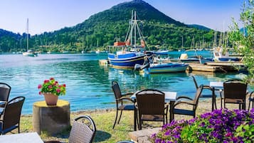 Un restaurante frente al agua con terraza, barcos amarrados cerca y una montaña al fondo.