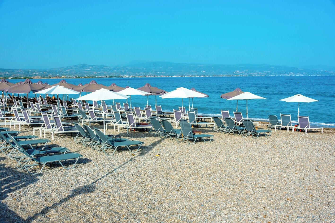 A beach with rows of sun loungers and umbrellas, a pebble beach, and the sea in the background.