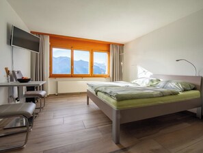 A hotel room with a bed, a desk with a chair, a TV, and a window with a mountain view. - Zurschmitten 8 2. UG Süd Studio, Dusche, WC 1 Zimmerwohnung, 2-Betten by Interhome (Riederalp)