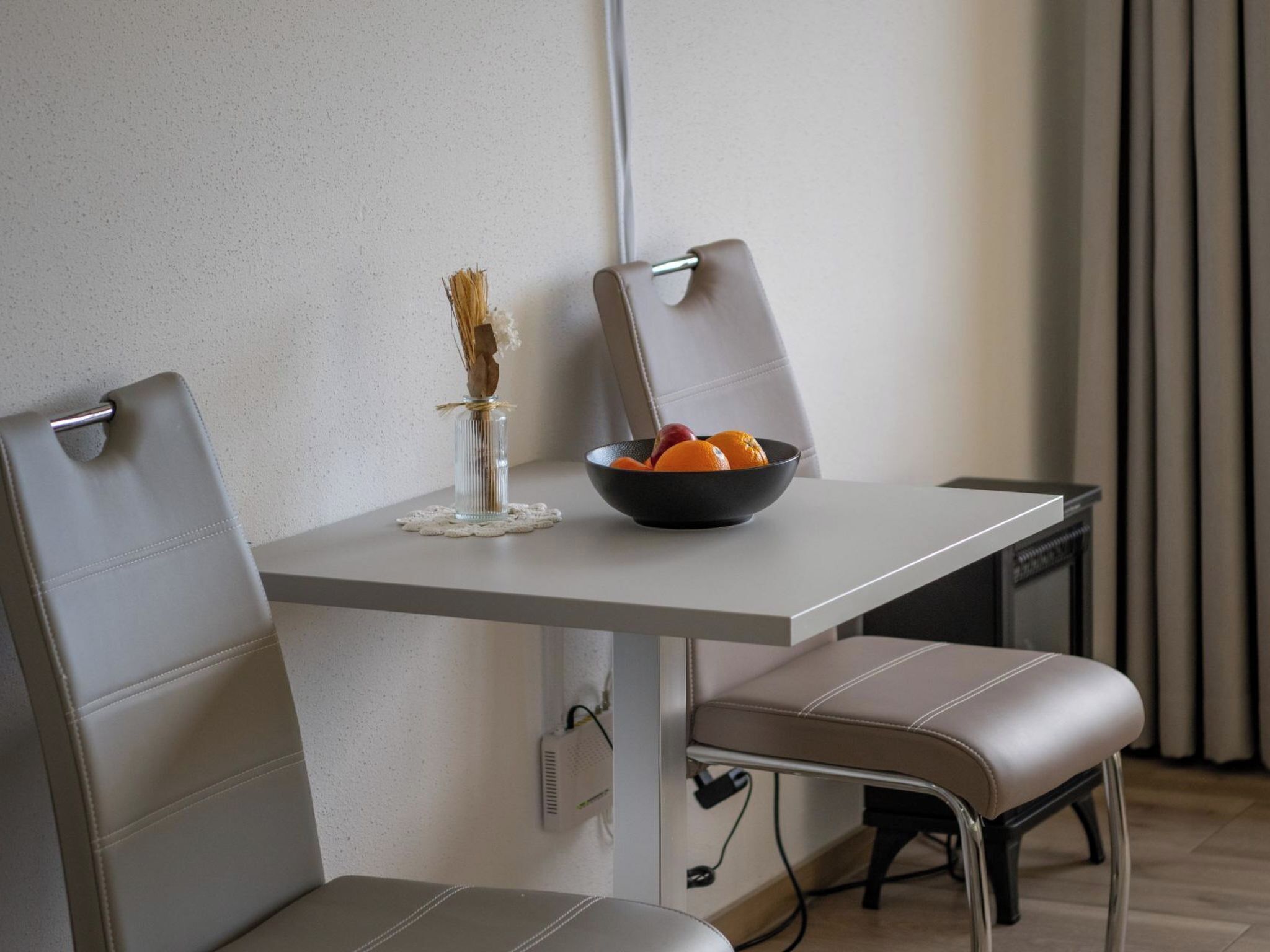 A modern dining area with a white table, two grey upholstered chairs, a black bowl with oranges, and a clear glass vase with dried plants.