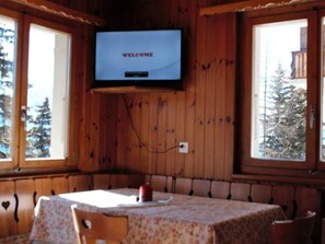 Interior d'una cabana de fusta amb una televisió que mostra 'WELCOME' a la pantalla, una taula de dinar amb cadires i vista a arbres a l'exterior.