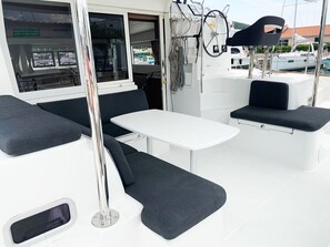 L'intérieur d'un bateau, avec une table blanche, deux coins salon dotés de sièges confortables aux couleurs sombres, et une fenêtre offrant une vue sur les bateaux et les bâtiments.
