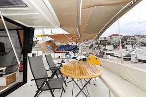 Cubierta de barco con mesa y sillas, un extintor y vistas a un puerto deportivo con otros barcos y edificios.