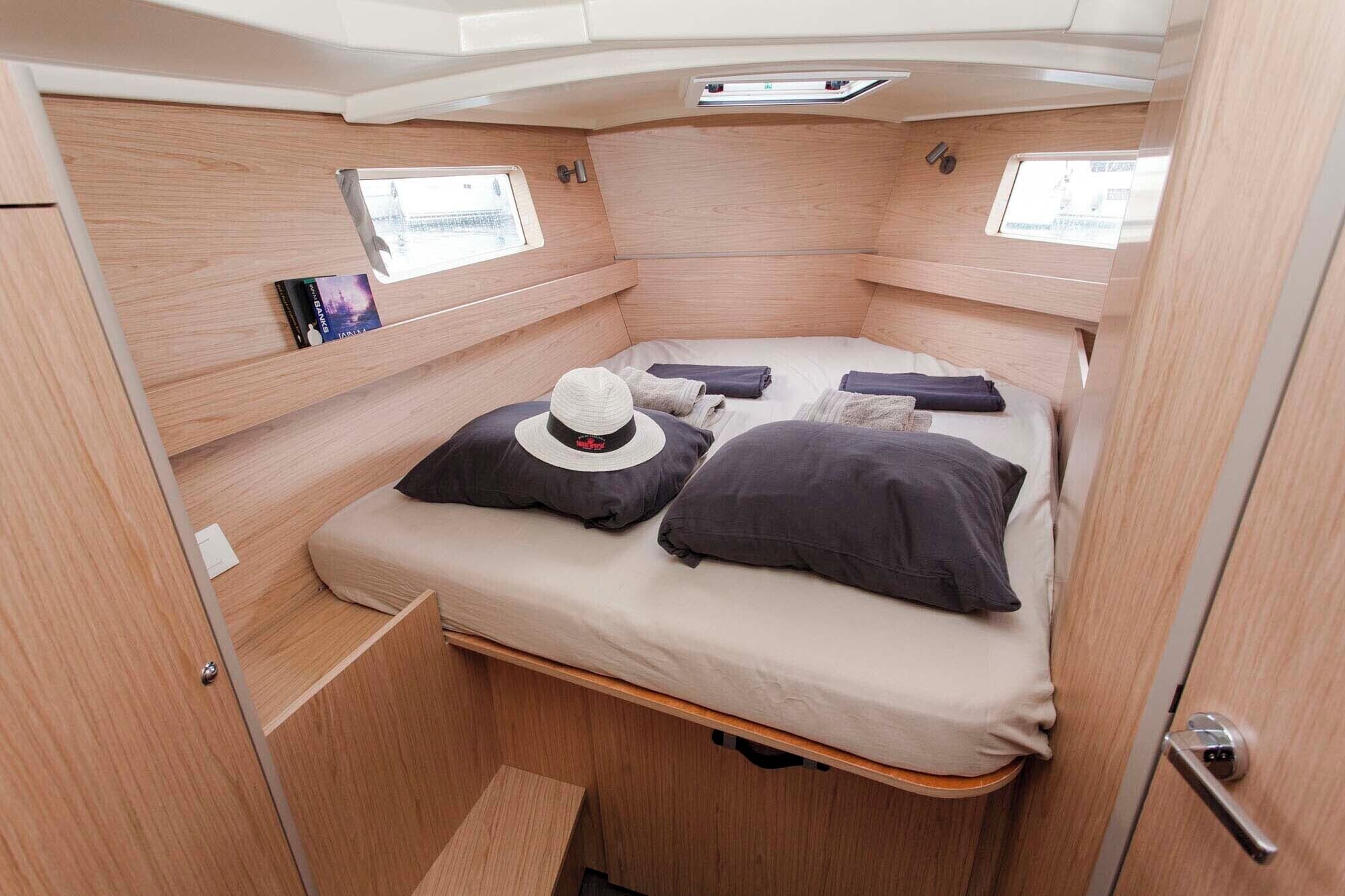 A compact, well-lit room with a bed, pillows, a hat, and a small window.
