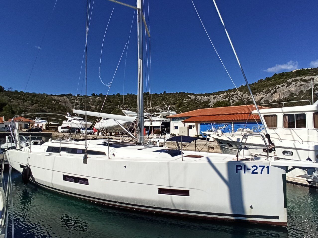 A marina with boats docked, a building with a red roof, and a hilly landscape in the background.