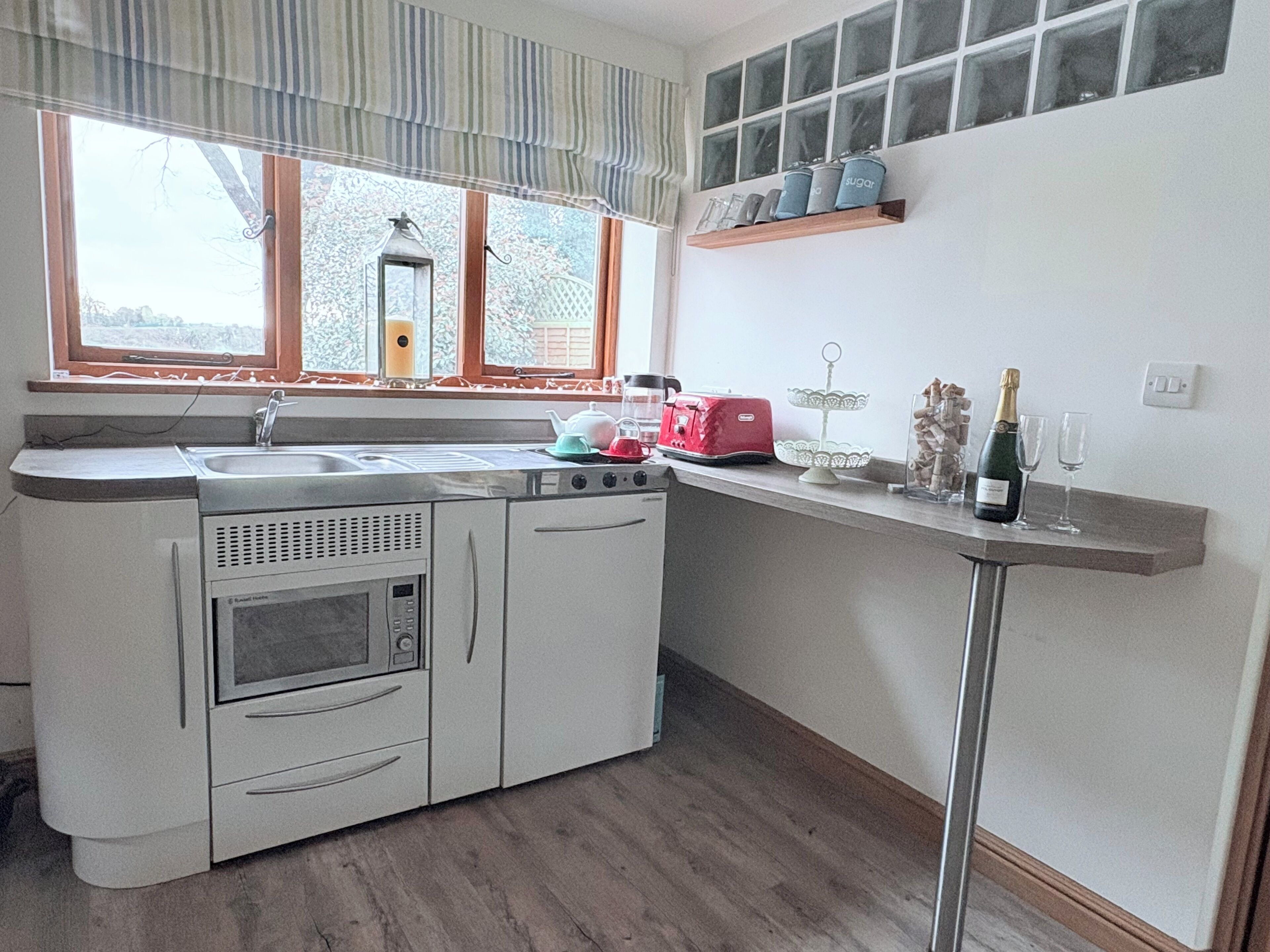 A compact kitchen with a sink, stove, and oven, a window with curtains, and a shelf with kitchen items.