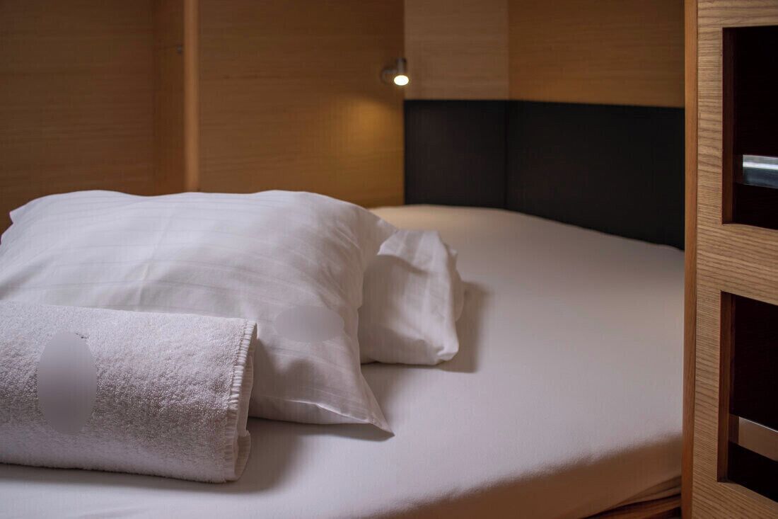 Un letto rifatto con lenzuola bianche e un asciugamano piegato, una testiera e un portalampada a parete.