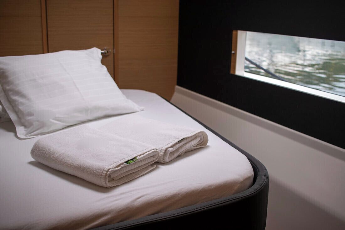 Un lit bien fait, avec un oreiller blanc et des serviettes pliées, une fenêtre donnant sur des arbres et une tête de lit en bois.