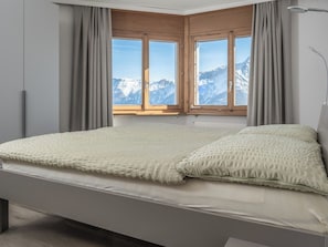 Un dormitorio con una cama, vistas de montañas nevadas y un ventanal con contraventanas de madera.