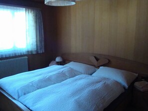 Ein Schlafzimmer mit einem hölzernen Kopfteil, einem Bett mit weißer Bettwäsche, einer Nachttischlampe und einem Fenster mit Vorhängen.
