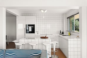 A modern kitchen with white cabinetry, a built-in oven, a dining area with bar stools, and a window providing a view of greenery. - Oceanfront Bondi Escape with Spectacular Views (Bondi)
