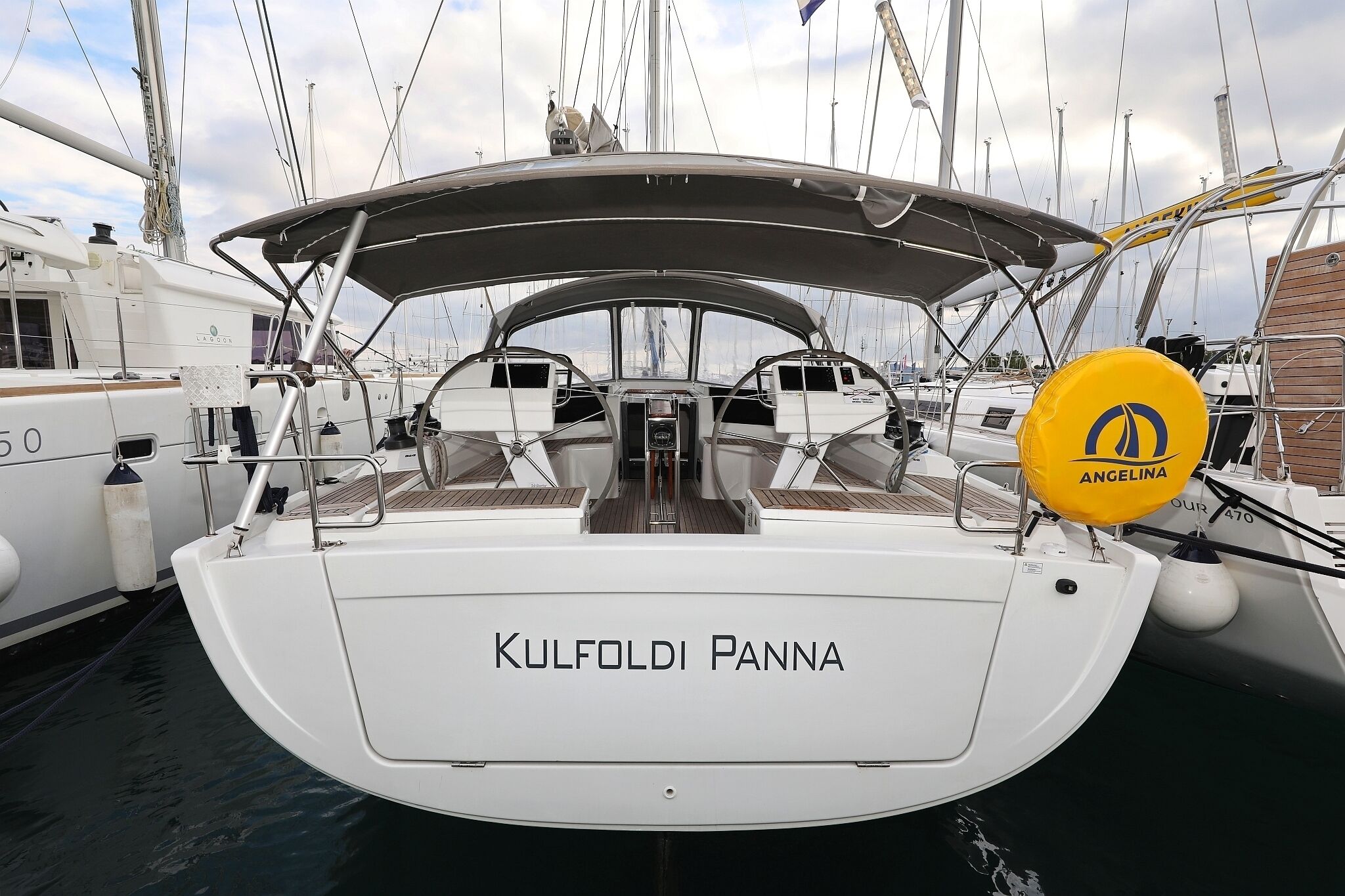 A white sailboat named 'KULFOLDI PANNA' docked at a marina with other boats, a covered deck, and a visible winch.