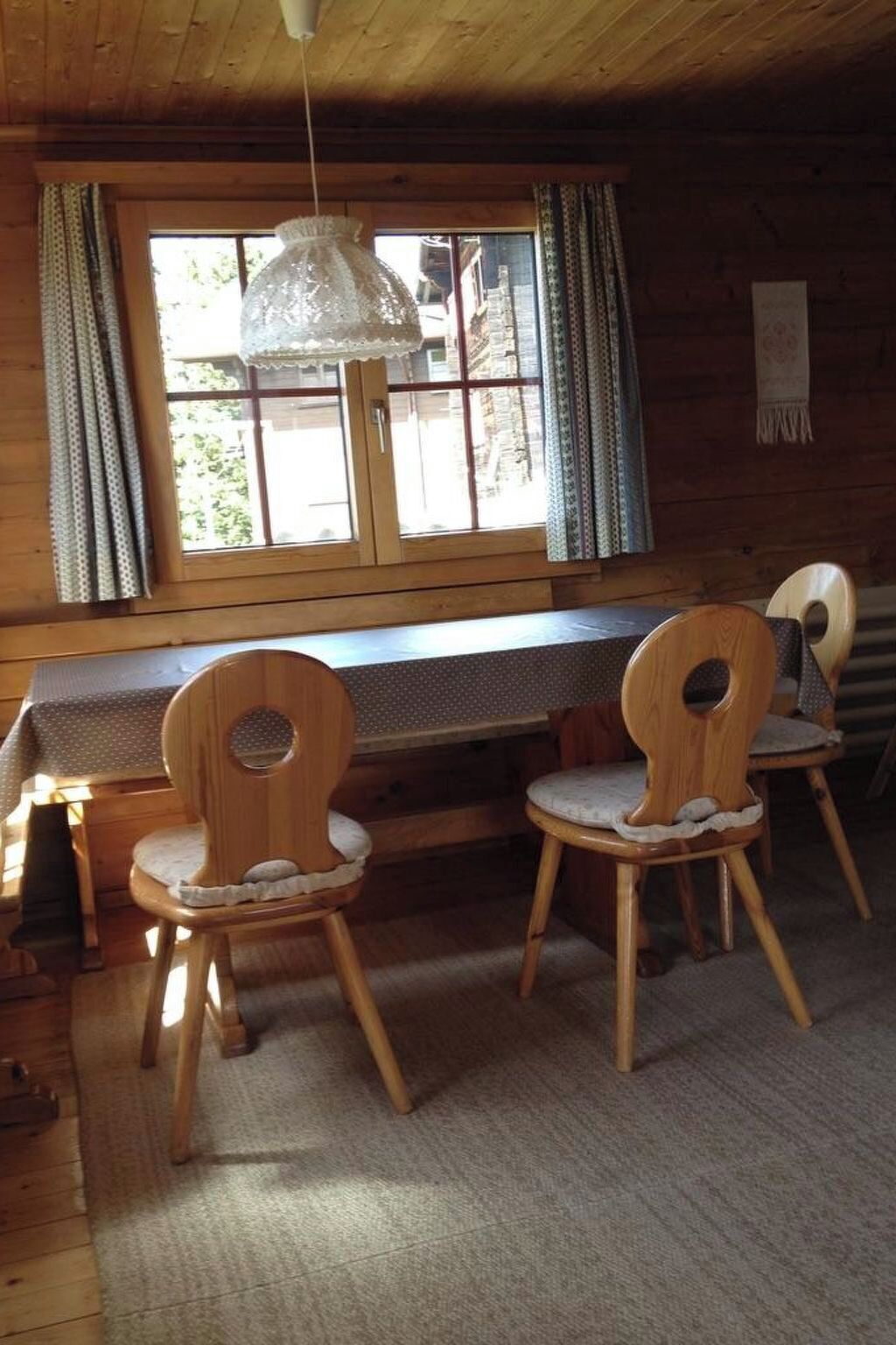 Interno di una baita in legno con un tavolo da pranzo e due sedie, una finestra con tende e un lampadario.