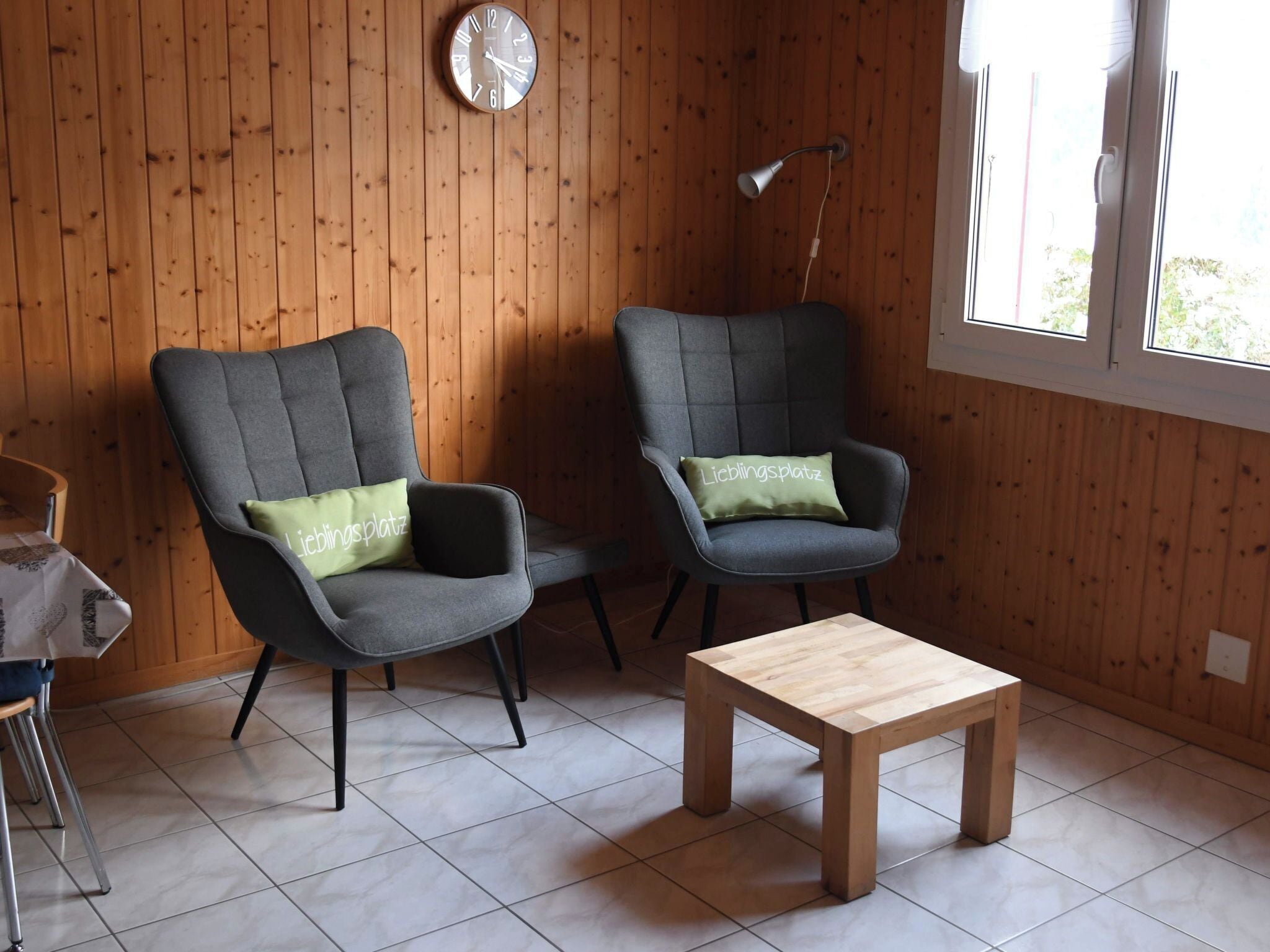 Una stanza con due poltrone grigie, un tavolino in legno, una sveglia appesa al muro e una finestra vista.
