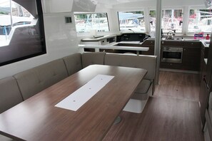 A boat interior with a dining table, cushioned seating, a kitchen area, and windows with a marina view. - Dubrovnik Delight: A Sun-Kissed Sailing Experience (Komolac)