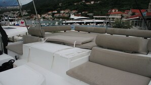 A boat deck with seating areas, a white cooler, and a view of a marina with houses in the background. - Dubrovnik Delight: A Sun-Kissed Sailing Experience (Komolac)