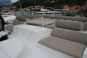 A boat deck with seating areas, a white cooler, and a view of a marina with houses in the background. - Dubrovnik Delight: A Sun-Kissed Sailing Experience (Komolac)