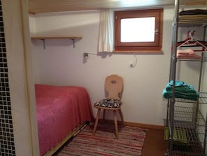 Ein kleines Zimmer mit einem Einzelbett, einem Sessel, einem Fenster und einem Metallregal mit zusammengelegter Kleidung.