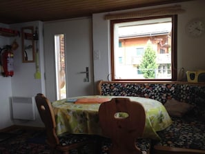 Ein Zimmer mit Esstisch, Stühlen, einem Fenster mit Blick auf Gebäude, einem Feuerlöscher und einer Wanduhr.