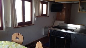 Ett litet kök med en spis, ett litet matbord och stolar. Det finns ett fönster med gardiner, en tv som är monterad på väggen och ett element.