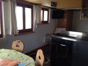 Eine kompakte Küche mit Herd, einem kleinen Esstisch und Stühlen. Ein Fenster mit Vorhängen, ein an der Wand befestigter Fernseher und eine Heizung sind ebenfalls vorhanden.