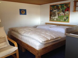 Ein Bett mit Holzrahmen und weißer Bettwäsche, ein Stuhl mit Streifenmuster-Kissen und ein Bild an der Wand.