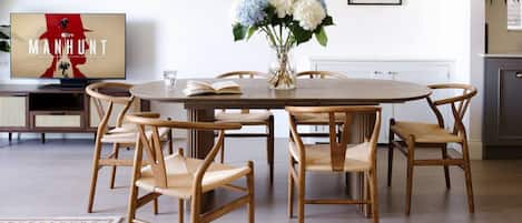 A dining area with a round table, six wooden chairs, a vase with flowers, a framed picture on the wall, and a flat-screen TV displaying a movie poster.