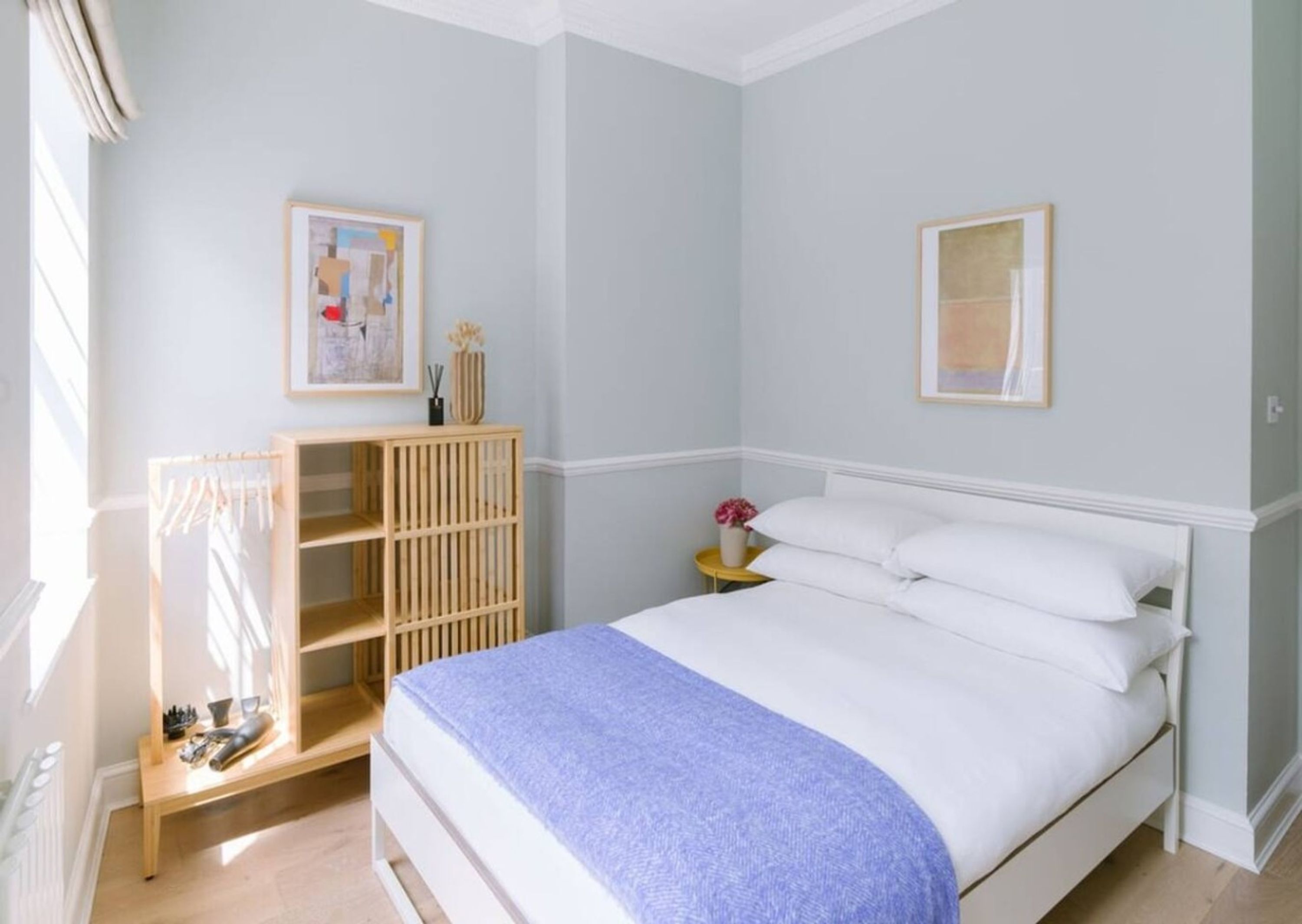 A bedroom with a bed, a wooden wardrobe, a nightstand with a vase, and two framed pictures on the wall.