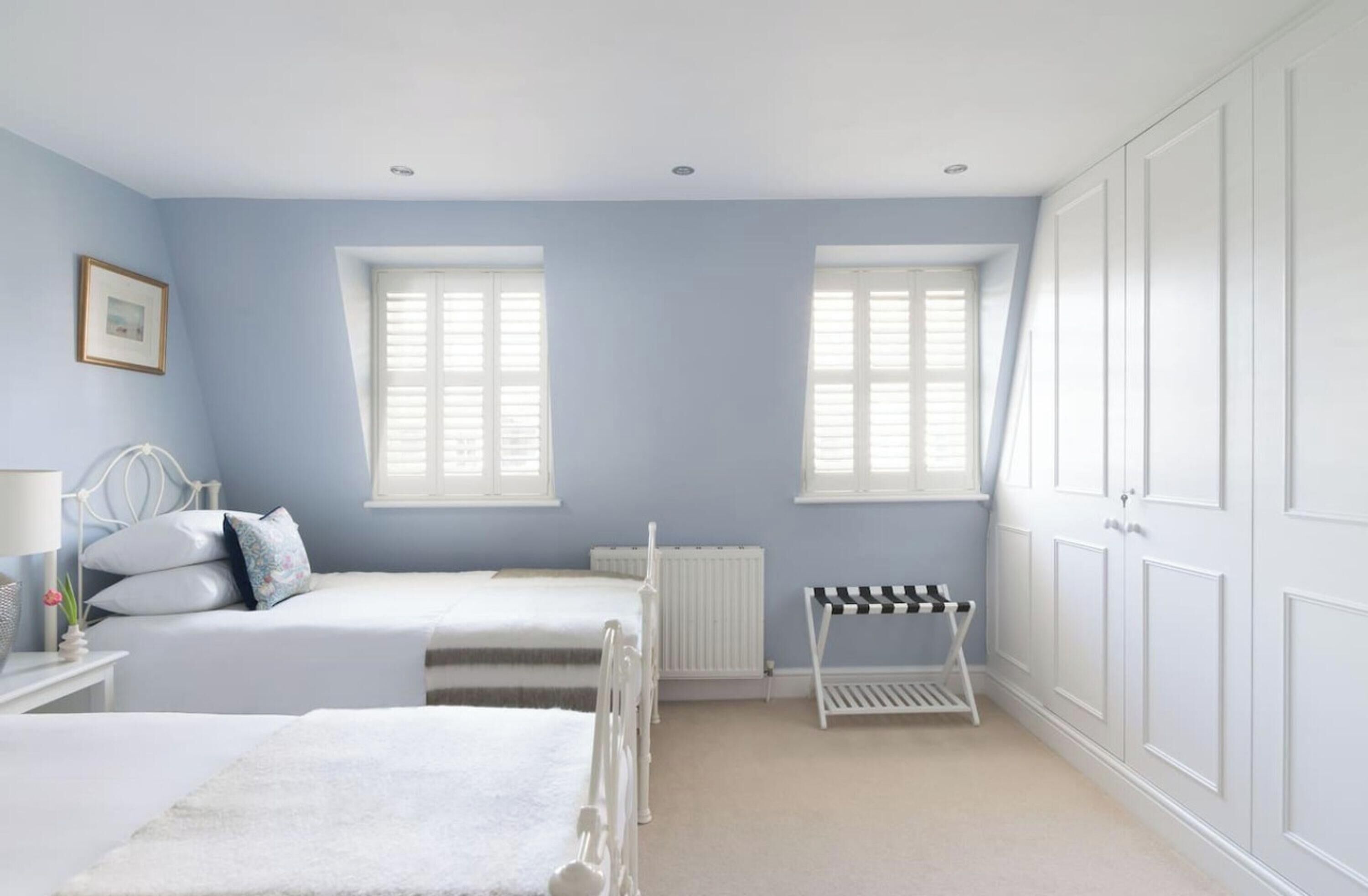 A bedroom with two beds, a radiator, a white radiator cover, a white bedside table with a lamp, a framed picture, and a window with shutters.