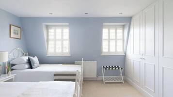 A bedroom with two beds, a radiator, a white radiator cover, a white bedside table with a lamp, a framed picture, and a window with shutters.