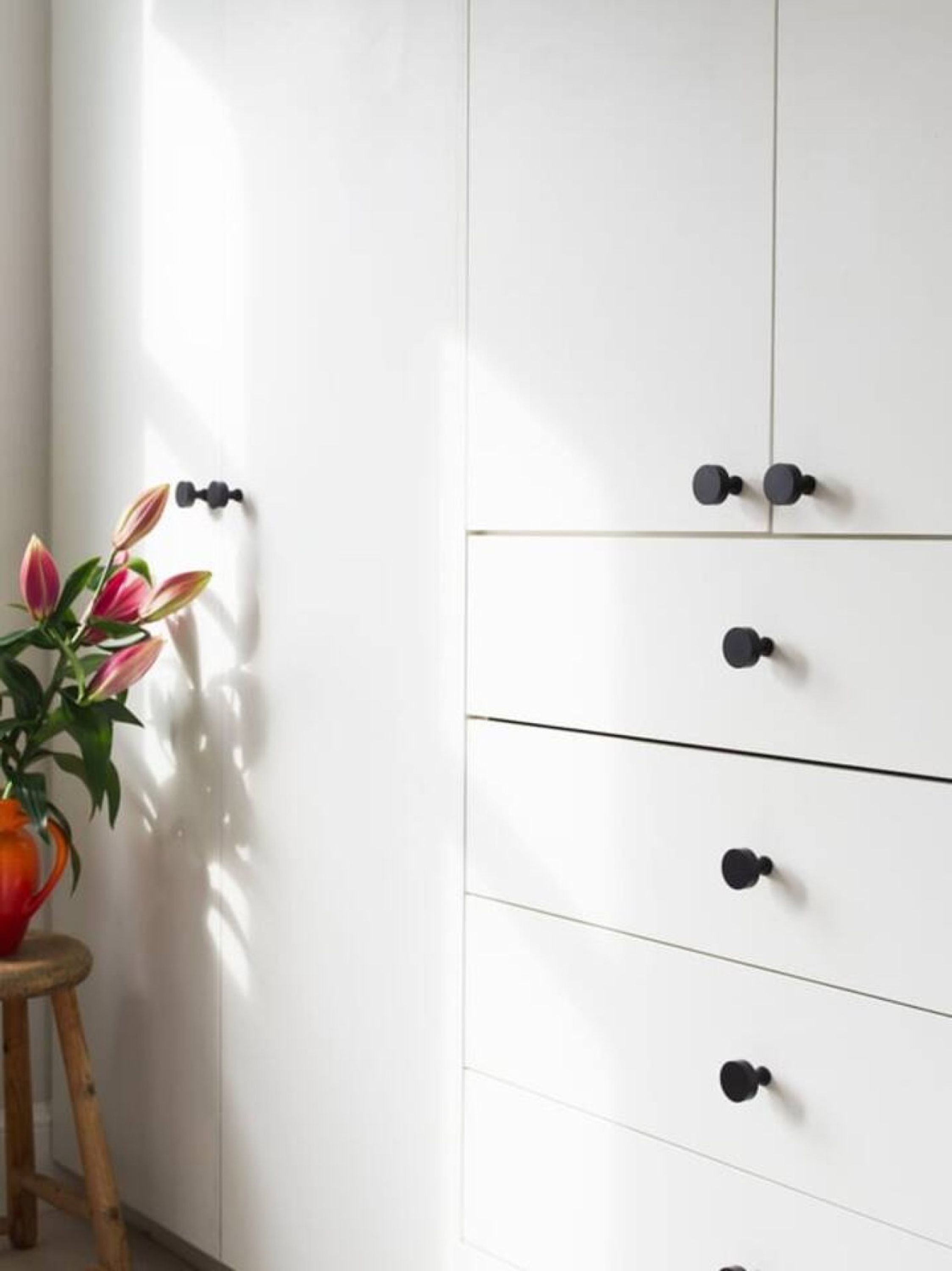 A white wardrobe with black handles, a wooden stool, and a potted plant with pink leaves.