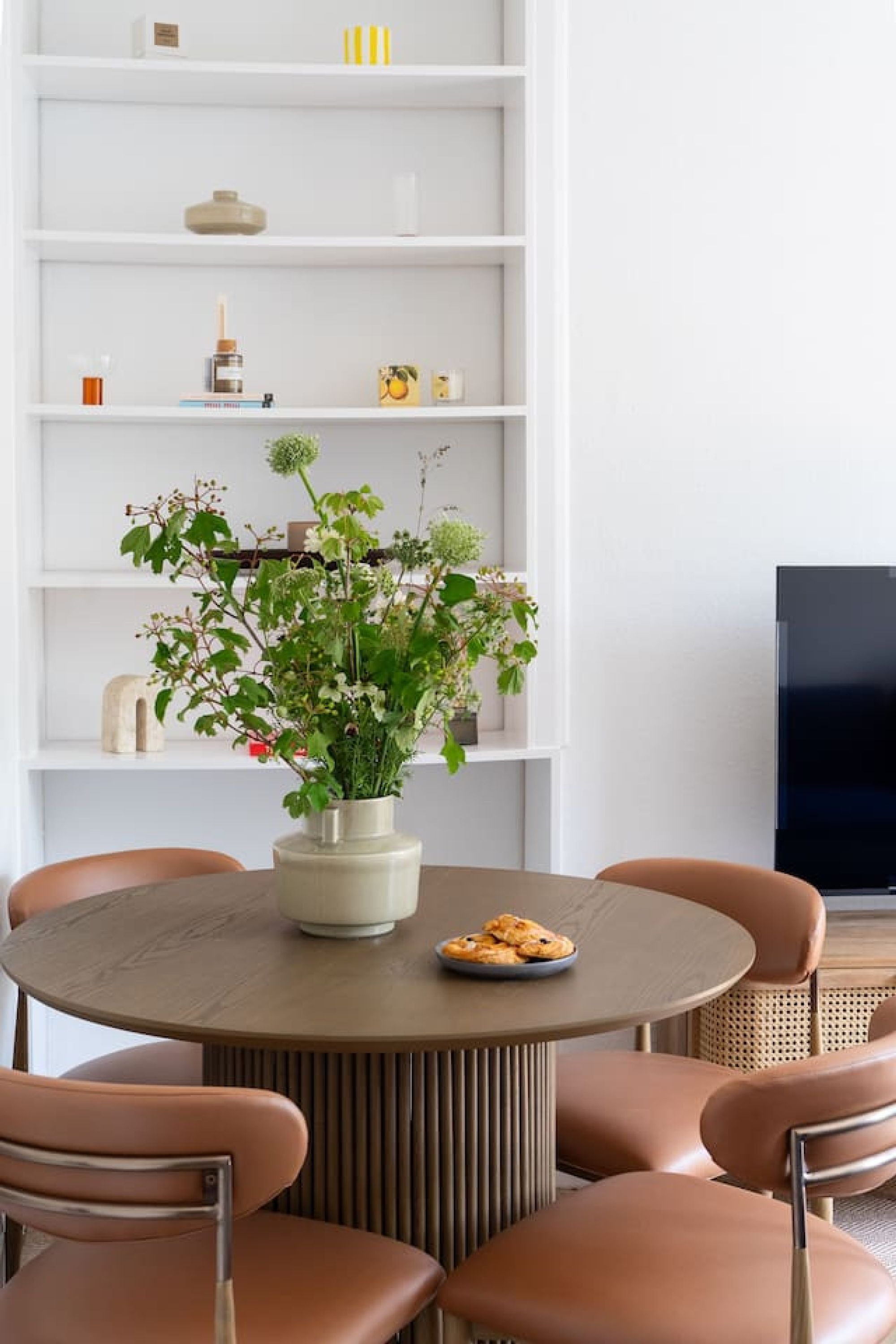 A round dining table with six chairs, a vase with flowers, and a plate of cookies.