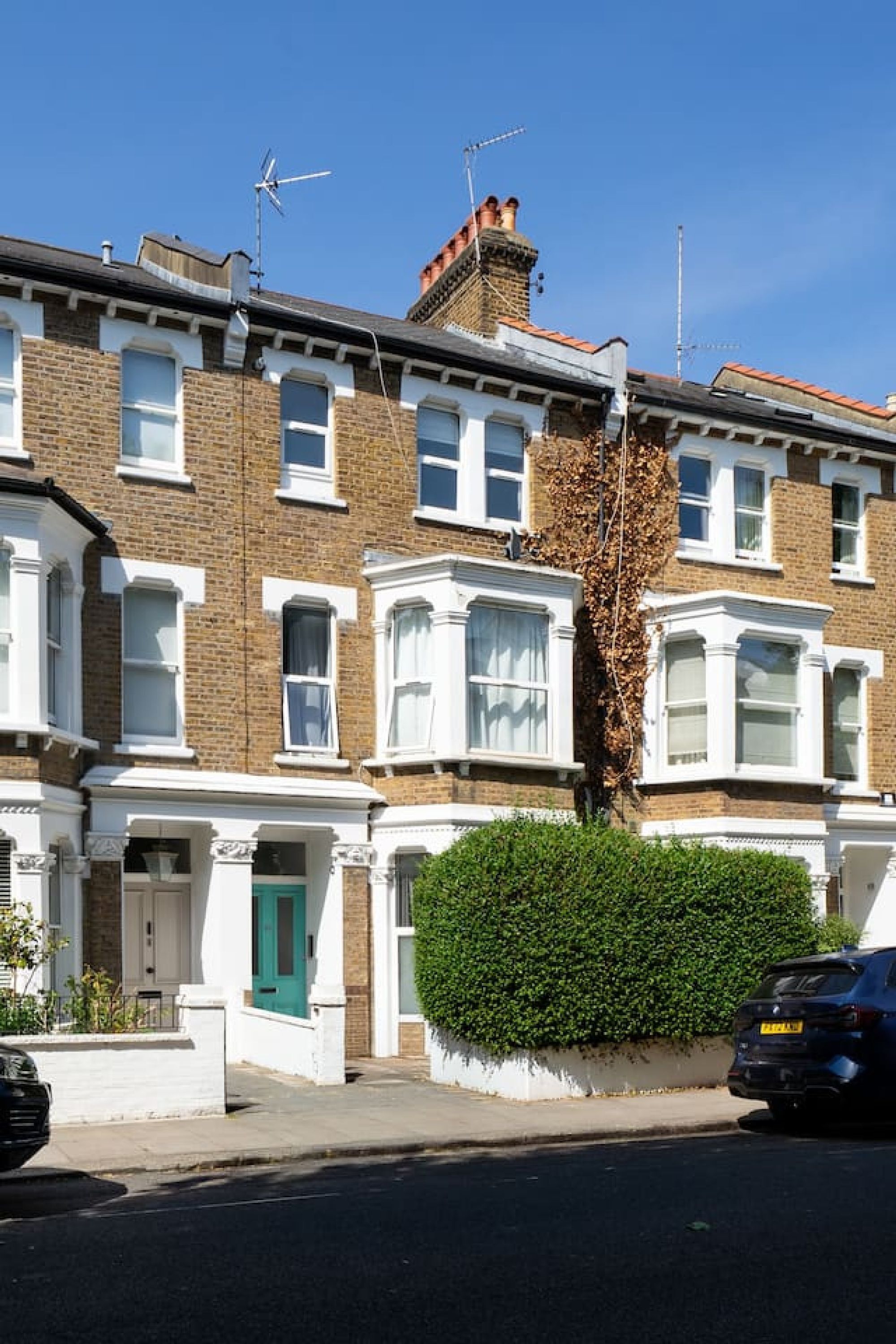 A row of terraced houses with a green door, a hedge, and a parked car.