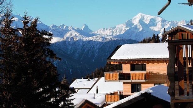 Eine Holzhütte mit Balkon, vor einer schneebedeckten Berglandschaft und blauem Himmel.
