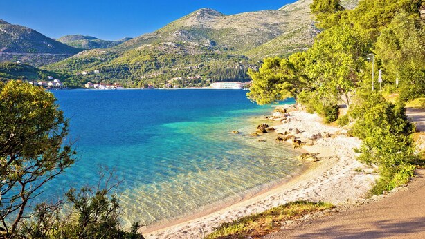 Paisatge costaner amb aigua cristal·lina, una platja sorrenca i muntanyes al fons.