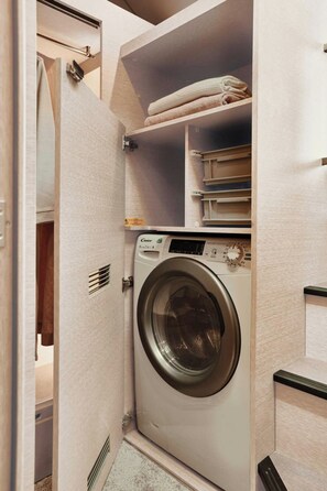 A compact laundry room with a front-loading washing machine, shelves for towels, and a small storage area. - Sun-kissed Sailing Adventure in Split-Dalmatia, Croatia (Trogir)