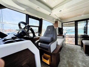 A boat's interior with a steering wheel, a cushioned seat, a small table, and a view of the marina through large windows. - Sun-kissed Sailing Adventure in Split-Dalmatia, Croatia (Trogir)