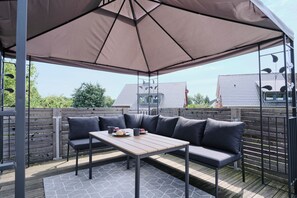 Un patio exterior con una mesa de madera, asientos acolchados y un toldo grande.