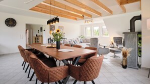 Un vaste espace de salle à manger avec une table en bois et des chaises marron, une cuisine moderne dotée d’une cuisinière intégrée, et un coin salon confortable avec un canapé gris et des fauteuils.