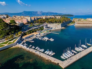 Vue aérienne d’une ville côtière avec un port de plaisance, des bâtiments et une chaîne de montagnes en arrière-plan.