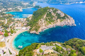 Vue aérienne d’une ville côtière, avec une imposante île rocheuse, des eaux d’un bleu limpide et une plage où se trouvent des bateaux et un stade.