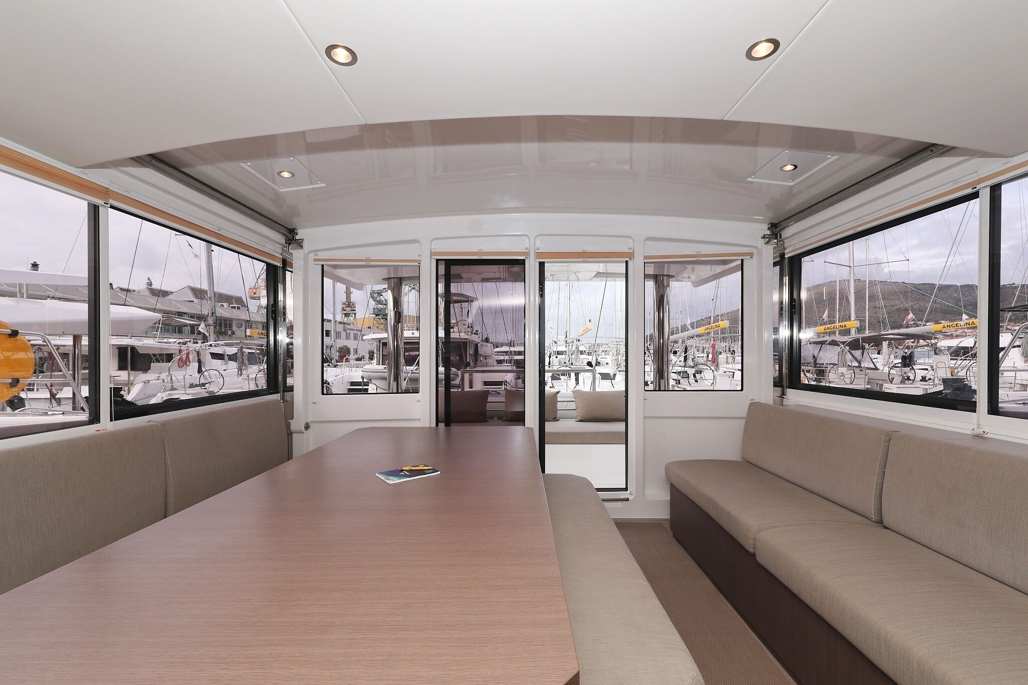 A modern interior with a long table, cushioned seating, large windows, and a view of boats outside.