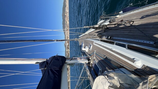 A sailboat's deck with multiple masts and rigging, a clear view of the coastline, and a calm sea.
