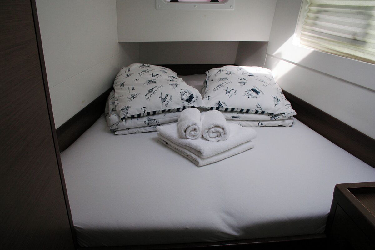 Un petit lit bien fait, avec des draps blancs et des oreillers à motifs, une serviette pliée, et une fenêtre avec des stores.